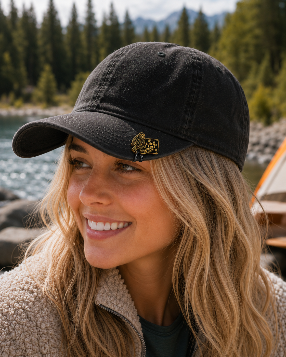 Blonde woman wearing a Bigfoot Knower Hat Clip on a black camping hat near a mountain lake